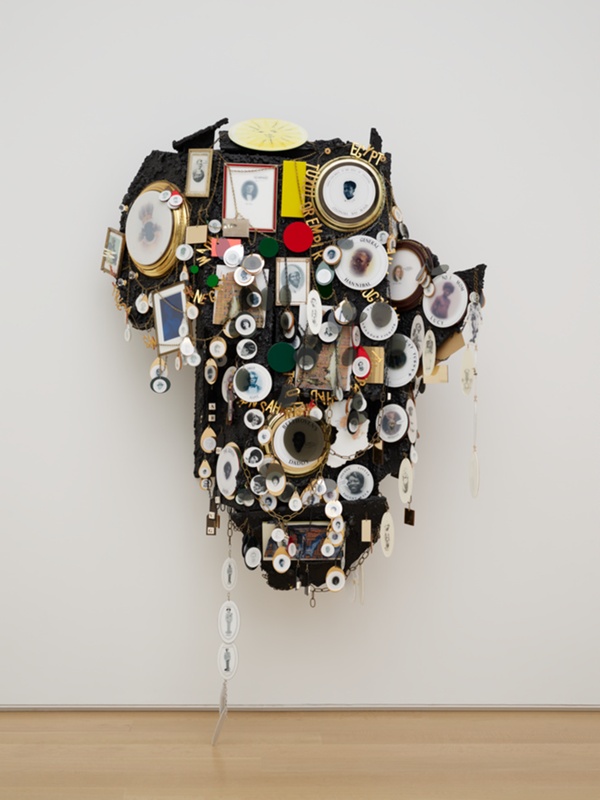 Large black sculpture in the shape of Africa. The form is covered in gold chains, circular photographs or "medallions" depicting prominent African American figures, and other images and text. The sculpture hangs on a white wall. 
"